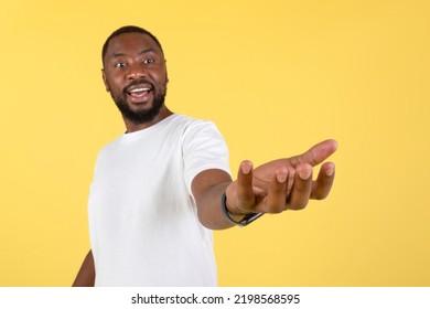 Excited African American Man Holding Invisible Object Showing And Advertising Your Product Posing Standing Over Yellow Studio Background, Wearing White T-Shirt. Shallow Depth