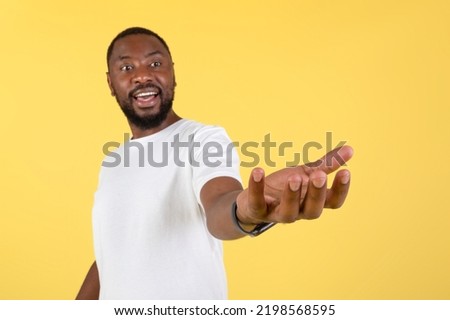 Excited African American Man Holding Invisible Object Showing And Advertising Your Product Posing Standing Over Yellow Studio Background, Wearing White T-Shirt. Shallow Depth