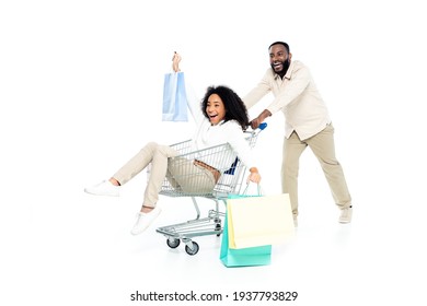 excited african american man moving shopping cart with happy wife and shopping bags on white