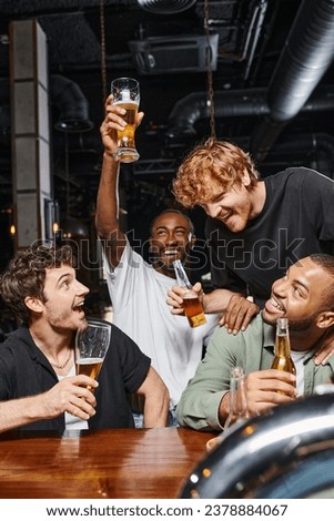 excited african american man raising glass of beer near cheerful friends at bar counter, nightlife
