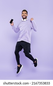 Excited African American Man With Wireless Headphones Holding Smartphone And Jumping On Purple