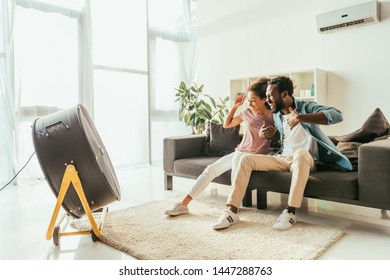 Excited African American Man And Woman Sitting On Sofa In Front Of Blowing Electric Fan