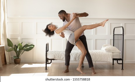 Excited African American Millennial Husband And Wife Dance Swirl In Bedroom, Happy Biracial Mixed Race Dancer Couple In Pajamas Have Fun Swaying Together At Home. Relocation, Relationship Goal Concept