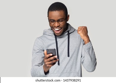 Excited african American millennial male in glasses isolated on grey studio background hold smartphone happy wining online, overjoyed black man look at cellphone smile feel joyful reading good news