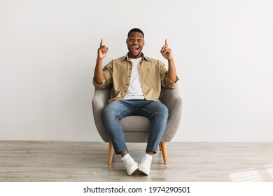 Excited African American Millennial Man Pointing Fingers Up Sitting In Chair Over Gray Wall Background, Looking At Camera. Check This Our, I Have Great Idea Concept. Front View - Shutterstock ID 1974290501