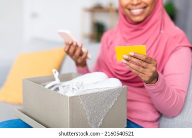 Excited african american muslim lady holding credit card and cellphone, unpacking cardboard box with new shoes, ordering and paying online from home