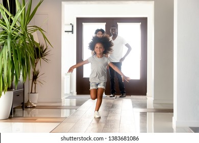 Excited African American Small Girl Run Into New Home Relocating With Young Parents, Happy Family Moving To Own House, Little Daughter Have Fun Jumping In Hallway. Ownership Concept