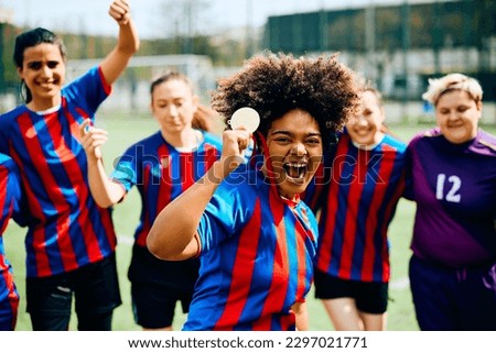 Excited African American soccer player with gold medal celebrating with her teammates on playing field and looking at camera. 