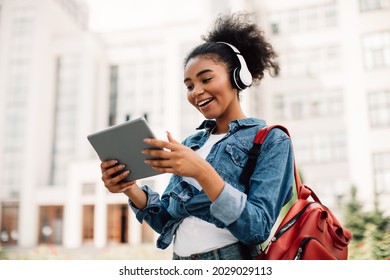 Excited African American Student Girl Using Digital Tablet Wearing Headphones Standing Near University Building Outdoors. Educational Website, E-Learning Concept. People And Gadgets - Powered by Shutterstock