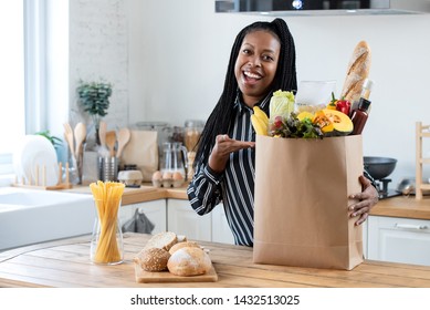 Excited African American Woman With Grocery Paper Bag Filled With Fresh Vegetables And Bread On Kitchen Platform Looking At Camera At Home