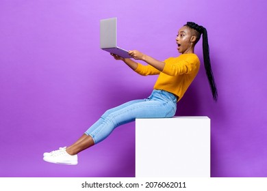 Excited African American woman holding and watching  laptop computer while sitting on stool in isolated purple color studio background - Shutterstock ID 2076062011