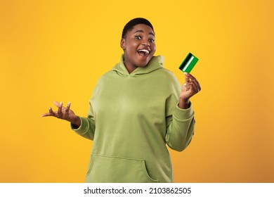 Excited African American Woman Holding Credit Card In Hand Showing It To Camera And Smiling Standing Over Yellow Studio Background. Finances And Bank Service, Easy Payment Concept