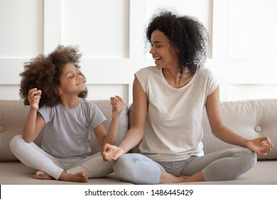 Excited african american woman teaching cute little adorable daughter meditating and practicing yoga exercises. Happy mixed race family sitting on comfy couch in lotus position, having fun together. - Powered by Shutterstock