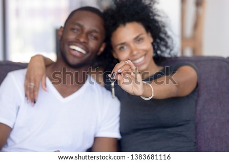 Excited African American young family show keys to own home, happy black couple sit on couch praise buying first house together, smiling husband and wife purchase new property. Ownership concept