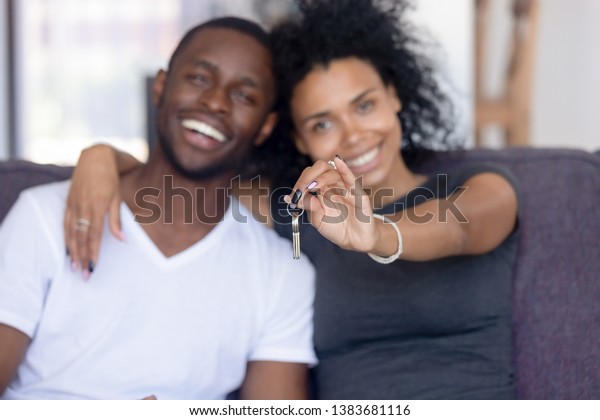 Excited African American young family show keys\
to own home, happy black couple sit on couch praise buying first\
house together, smiling husband and wife purchase new property.\
Ownership concept