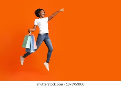 Excited African American Young Woman With Colorful Shopping Bags Jumping In The Air And Pointing At Copy Space For Sales Or Discounts Advertisement, Orange Studio Background, Panorama