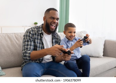 Excited African Dad And Son Playing Game Having Fun Sitting On Couch At Home. Young Black Father And His Kid Play Videogames Spending Time Together On Weekend Concept