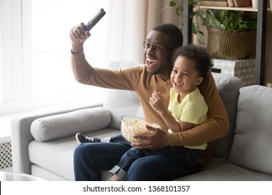 Excited african father raise hand hold remote control celebrate football team victory little son sitting on dad lap family eating popcorn watching sport program on tv, leisure activity at home concept