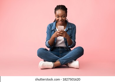 Excited African Lady With Cellphone Texting, Using Phone App And Browsing Internet Sitting Over Pink Background. Mobile Communication, Smartphone Application Concept. Empty Space, Studio Shot
