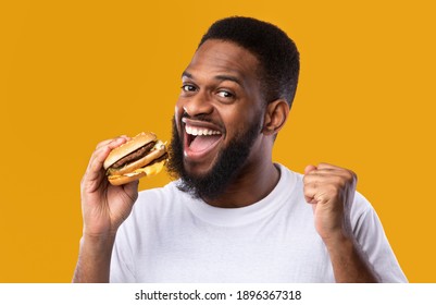 Excited African Man Eating Burger Gesturing Yes Enjoying Unhealthy Fast Food Standing Over Yellow Studio Background. Finally, Cheat Meal. Tasty Junk Food Concept