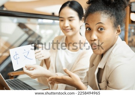 Excited African woman learning Chinese language with her female teacher, holding Chinese vocabulary word card reading 