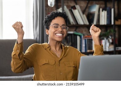 Excited African woman sit at desk with laptop scream with joy with clenched fists, celebrate great news, get scholarship approval, feels overjoyed looks happy. Moment of success, achievement concept