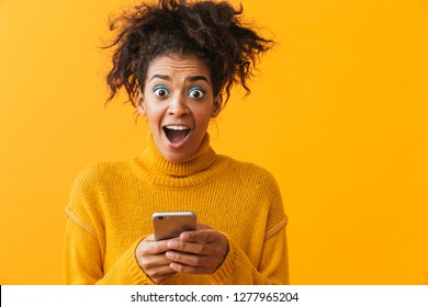 Excited African Woman Wearing Sweater Standing Isolated Over Yellow Background, Holding Mobile Phone