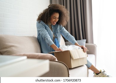 Excited african young woman shopper opening parcel box at home. Happy satisfied ethnic girl customer receiving online shop purchase by postal shipping, unpacking delivery receiving gift sit on sofa.