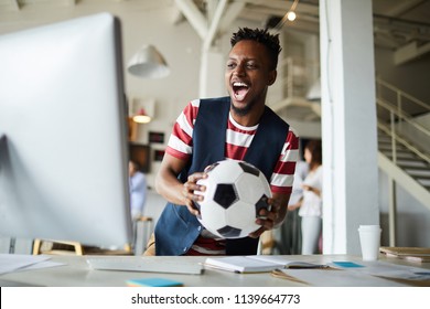 Excited African-american Guy With Soccer Ball Shouting In Front Of Computer Monitor While Watching Webcast