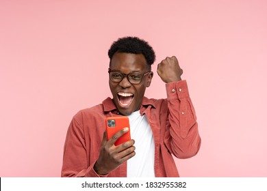 Excited Afro-American millennial man in glasses hold mobile phone isolated on pink studio background. Overjoyed black man look at smartphone, smile feel joyful reading good news, doing winner gesture.
