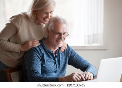 Excited Aged Husband Using Laptop Showing Smiling Wife Something On Screen, Happy Senior Couple Looking At Computer, Watching Pictures Together Or Funny Video Online. Elderly And Technology Concept