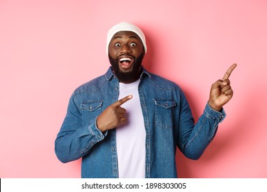 Excited And Amazed Black Man Showing Awesome Offer, Pointing Fingers Right At Copy Space, Standing In Hipster Beanie And Denim Shirt On Pink Background