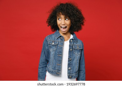 Excited Amazed Little African American Kid Girl 12-13 Years Old Wearing Casual Denim Jacket Looking Aside Isolated On Bright Red Color Background Children Studio Portrait. Childhood Lifestyle Concept