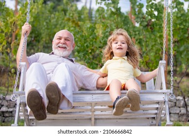 Excited Amazed Old Grandfather And Young Child Grandson Swinging In Garden Outdoors. Grand Dad And Grandson Sitting On Swing In Park.