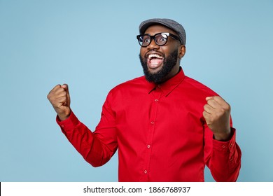 Excited amazed young bearded african american man 20s wearing casual red shirt cap eyeglasses standing doing winner gesture clenching fists isolated on pastel blue color background studio portrait