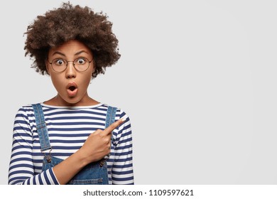 Excited anxious lovely young African female with curly black hair, dark skin, keeps mouth opened, stares with surprisement, points finger in side against white blank wall for your promotional text.