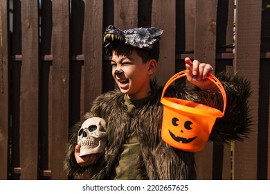 Excited Asian Boy In Werewolf Costume Holding Halloween Bucket And Spooky Skull While Looking Away