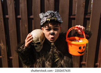 Excited Asian Boy In Werewolf Costume Holding Skull And Halloween Bucket With Sweets