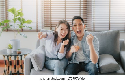 Excited Asian couple celebrating victory of sport competition on tv screen while sitting on sofa in living room at home