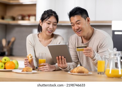 Excited Asian Couple Shopping From Home While Having Breakfast Together, Happy Chinese Middle Aged Man And Young Woman Sitting At Kitchen Table, Drinking Juice, Holding Digital Tablet And Credit Card