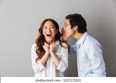 Excited asian couple standing isolated over gray background, telling secrets to each other