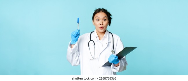 Excited Asian Female Physician Holding Clipboard, Raising Pen Up In Eureka Sign, Has Revelation, Pitching An Idea, Standing On Blue Background