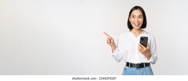 Excited Asian Girl Shopping On Mobile App, Holding Phone And Pointing Finger Left At Empty Copy Space, Showing Promo Text, Standing With Smartphone Against White Background