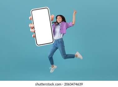 Excited asian lady showing empty smartphone screen while jumping up over blue studio background, collage, full body length, banner. Happy woman recommending cool mobile app or website