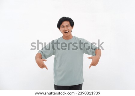 Excited Asian male employee wearing a T-shirt pointing at the copy space below him, isolated by a white background