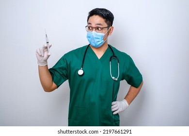 Excited Asian Male Nurse In Medical Mask, Rubber Gloves And Scrubs, Looking Amazed At Syringe With Vaccine, Isolated Over White Background