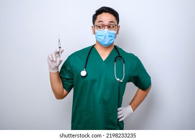 Excited Asian Male Nurse In Medical Mask, Rubber Gloves And Scrubs, Looking Amazed At Syringe With Vaccine, Isolated Over White Background