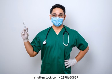 Excited Asian Male Nurse In Medical Mask, Rubber Gloves And Scrubs, Looking Amazed At Syringe With Vaccine, Isolated Over White Background