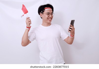 Excited Asian man celebrate Indonesian independence day by holding cell phone and flag. Isolated - Shutterstock ID 2283877015