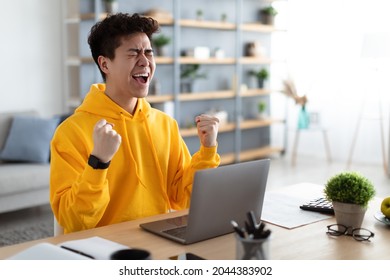Excited asian man emotionally celebrating success using laptop in home office, sitting at desk raising hands with joy, shaking fists, received good news, got promotion, finished project, free space - Shutterstock ID 2044383902